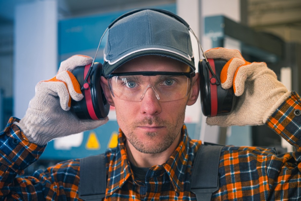 A man in safety glasses, gloves, and a plaid shirt adjusts earmuffs in an industrial setting. He looks directly at the camera, emphasizing noise protection and workplace safety.