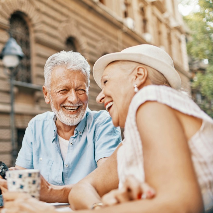 Two older adults, a man and a woman, sit outdoors at a table, smiling and enjoying each others company. The man has gray hair and a beard; the woman wears a light hat. They appear relaxed and happy.
