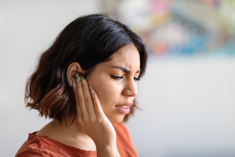 A woman with short brown hair touches her ear and looks uncomfortable, possibly experiencing ear pain, with a blurred colorful background behind her.